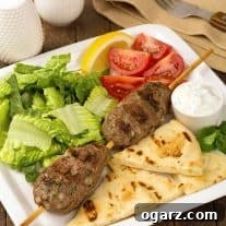 A dinner plate with two grilled skewered lamb meatballs (kofta), grilled pita bread, a small bowl of tzatziki sauce, lemon wedges, lettuce, and tomatoes; a fork, napkin, and salt and pepper shakers in the background.