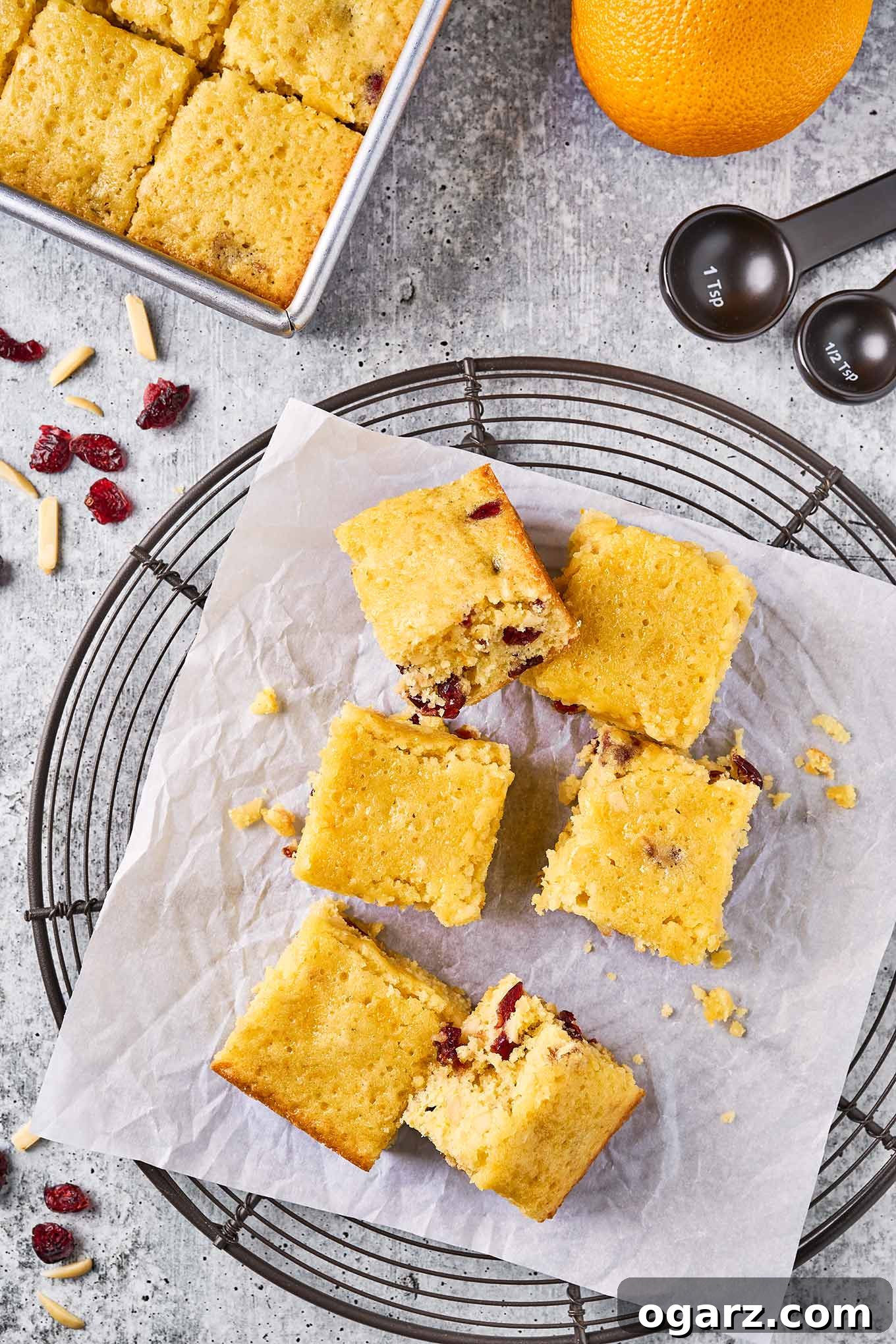 Zesty Cranberry Orange Squares 6 An overhead view of a round cooling rack with cranberry orange bars on a piece of parchment paper, surrounded by the baking pan and other ingredients.
