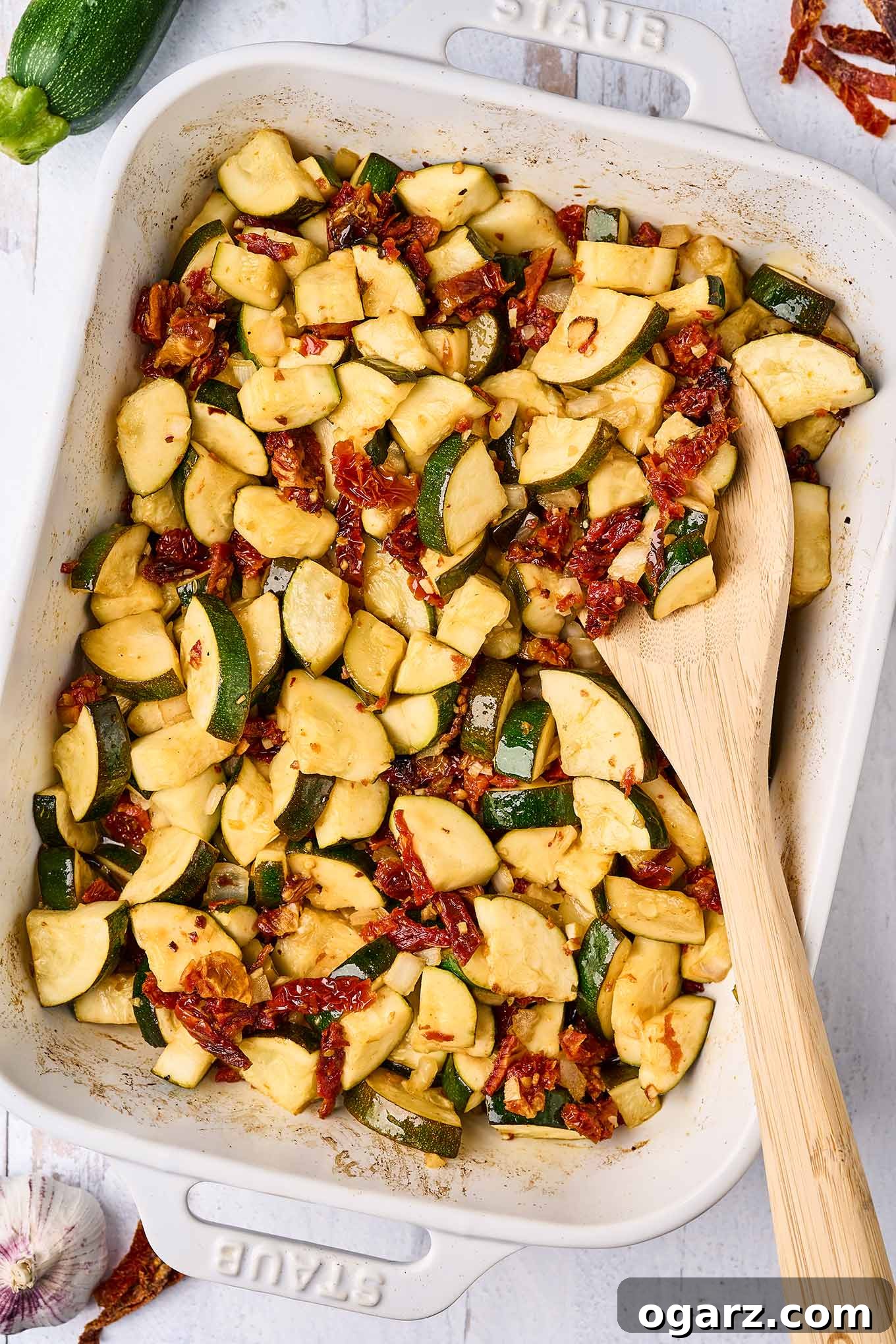 Sun-Kissed Zucchini & Garlic Bake 2 An overhead view of a casserole of baked zucchini chunks with garlic and sun-dried tomatoes, a wooden spoon in the dish and scattered sun-dried tomatoes, a bulb of garlic, and a whole zucchini surrounding it.