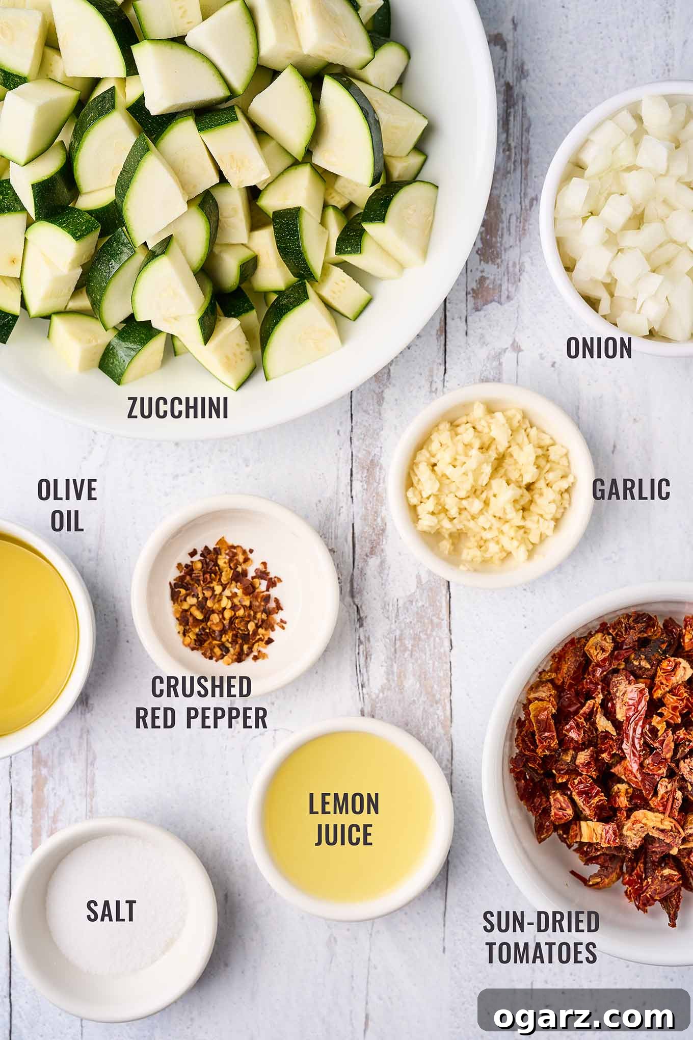 Sun-Kissed Zucchini & Garlic Bake 3 A labeled, overhead view of the ingredients needed to make a baked zucchini recipe; chunks of zucchini, sun-dried tomatoes, chopped onion, minced garlic, crushed red pepper, lemon juice, olive oil, and salt.