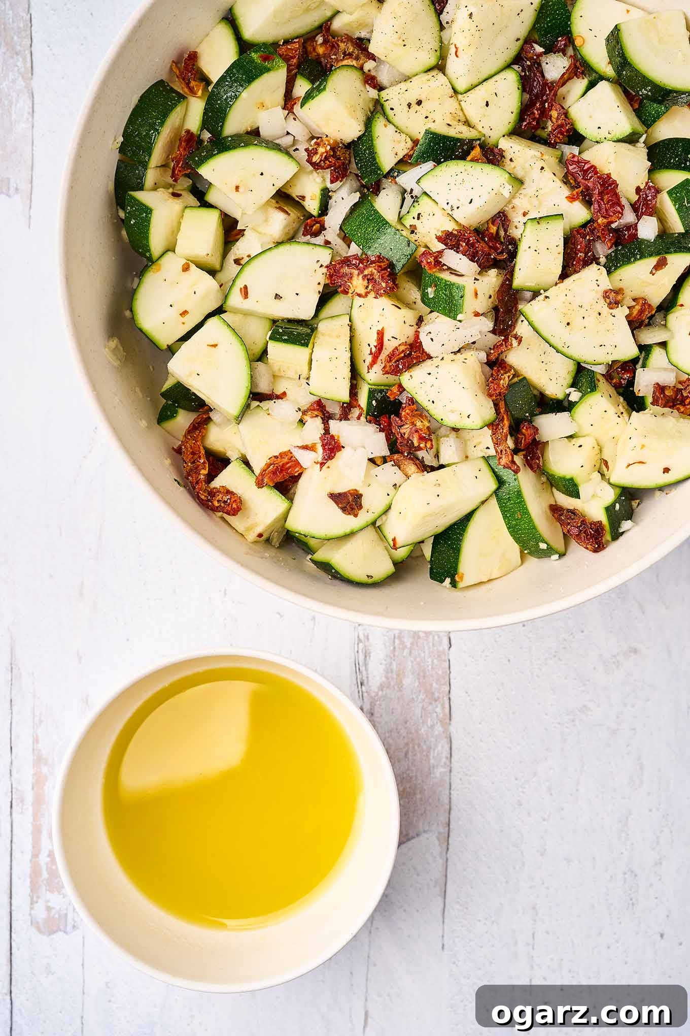 Sun-Kissed Zucchini & Garlic Bake 4 A bowl of seasoned chunks of zucchini, onion, garlic, and sun-dried tomatoes next to a dish of olive oil, ready to be combined and transferred to a casserole for baking.