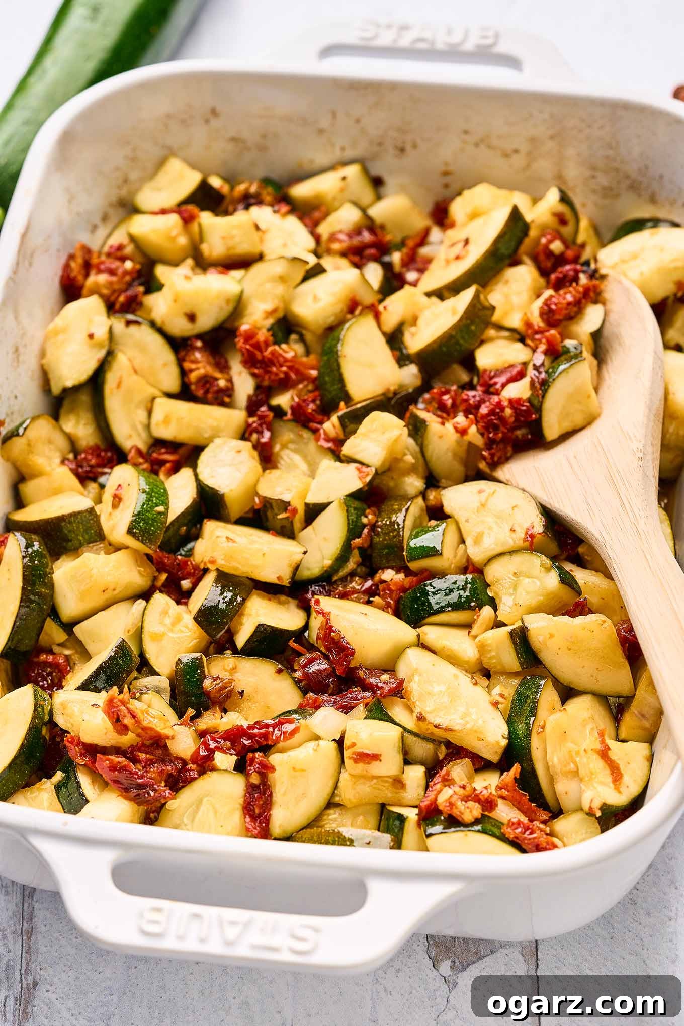 Sun-Kissed Zucchini & Garlic Bake 6 A close up view of a white, oblong casserole dish of baked zucchini with garlic and sun-dried tomatoes with a wooden spoon in it for serving.