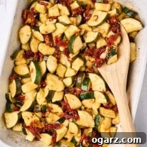 An overhead view of a casserole of baked zucchini chunks with garlic and sun-dried tomatoes, a wooden spoon in the dish and scattered sun-dried tomatoes, a bulb of garlic, and a whole zucchini surrounding it.