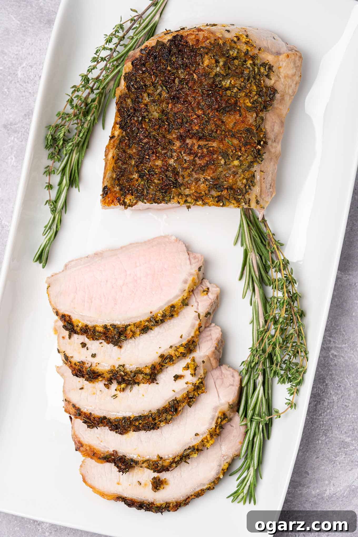 A partially carved, boneless pork loin roast crusted with garlic and fresh herbs on a serving platter, garnished with fresh rosemary and thyme.