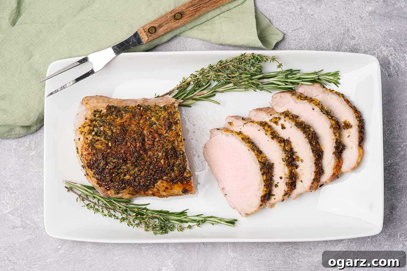 A partially sliced boneless pork roast on a serving platter with a carving fork, garnished with sprigs of fresh rosemary and thyme.