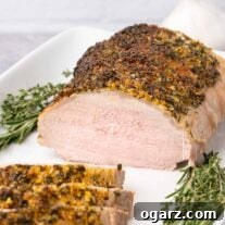 A partially carved, boneless pork loin roast crusted with garlic and fresh herbs on a serving platter, garnished with fresh rosemary and thyme.