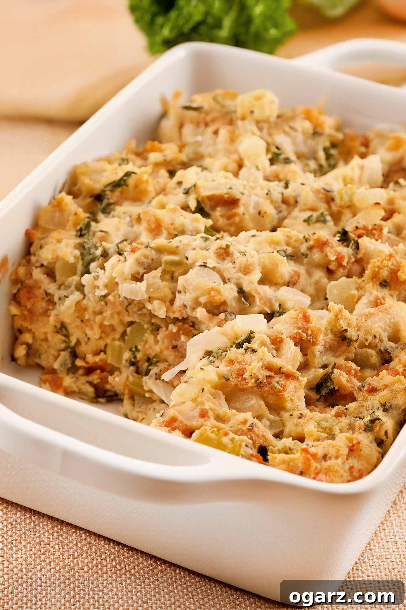 A golden-brown Italian sausage and herb bread stuffing in a white casserole dish, with a scoop removed, garnished with fresh celery leaves. Perfect for holiday feasts or a comforting family dinner.