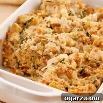 A white, oblong casserole dish filled of Italian sausage and herb bread stuffing with one serving removed, celery tops and a kitchen towel in the background.