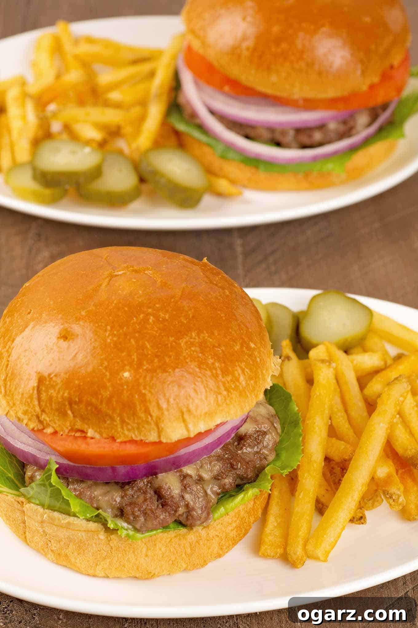 An overhead view of two plated servings of oven-baked cheeseburgers, served on soft buns with crisp lettuce, tomato, and onion. Each plate includes a generous portion of crispy French fries and tangy pickle slices on the side, creating a complete and satisfying meal.