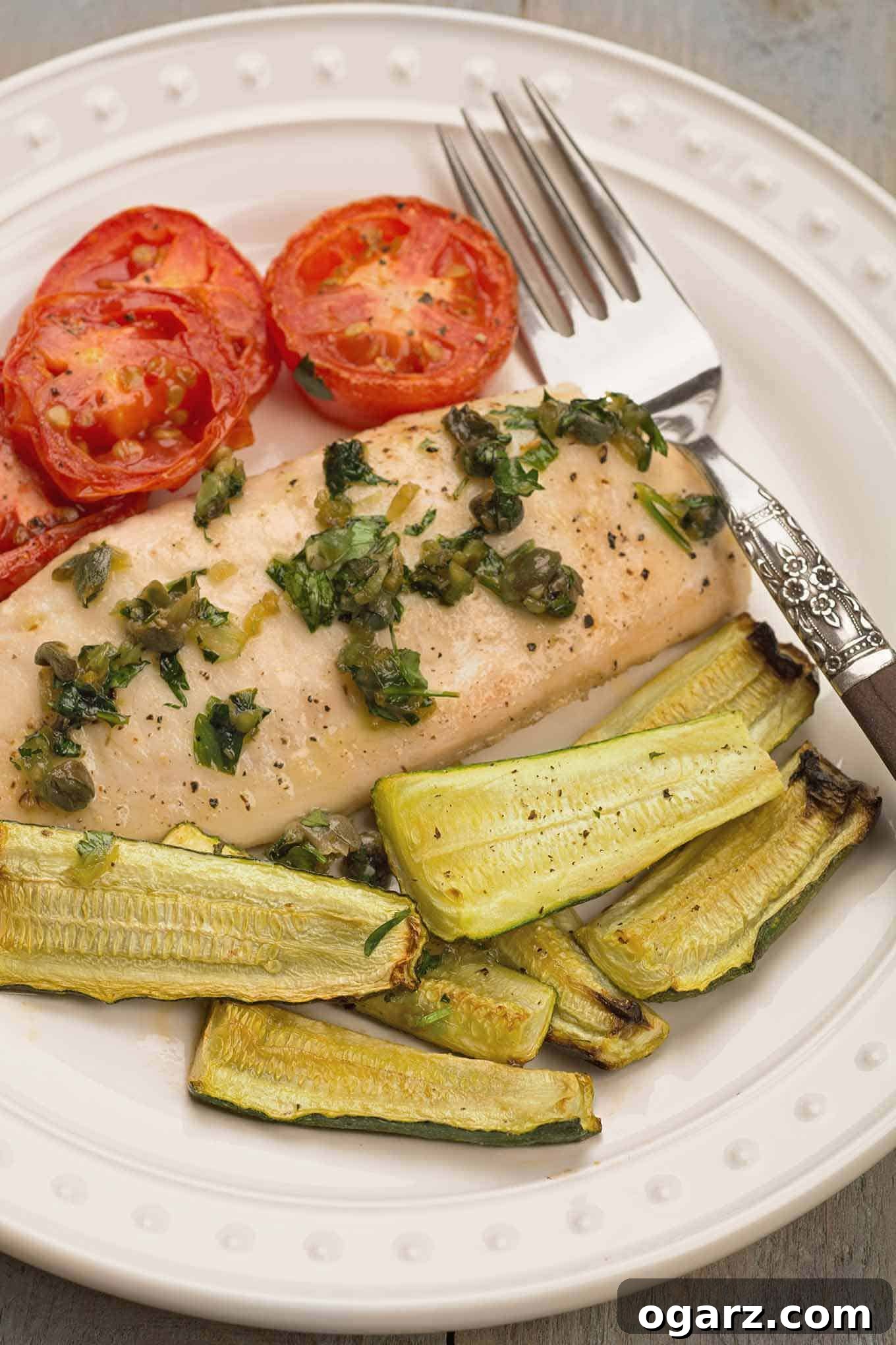One-Pan Mediterranean Fish Feast 3 A close-up view of a single serving plate, featuring a perfectly roasted tilapia fillet generously topped with a mixture of bright green capers and fresh parsley. Alongside are tender, caramelized plum tomato slices and vibrant zucchini wedges, creating a balanced and appetizing presentation.