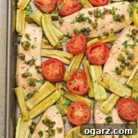 An overhead view of a Mediterranean-style fish dinner on a sheet pan, including tilapia fillets, sliced plum tomatoes, zucchini wedges, capers, and fresh parsley.