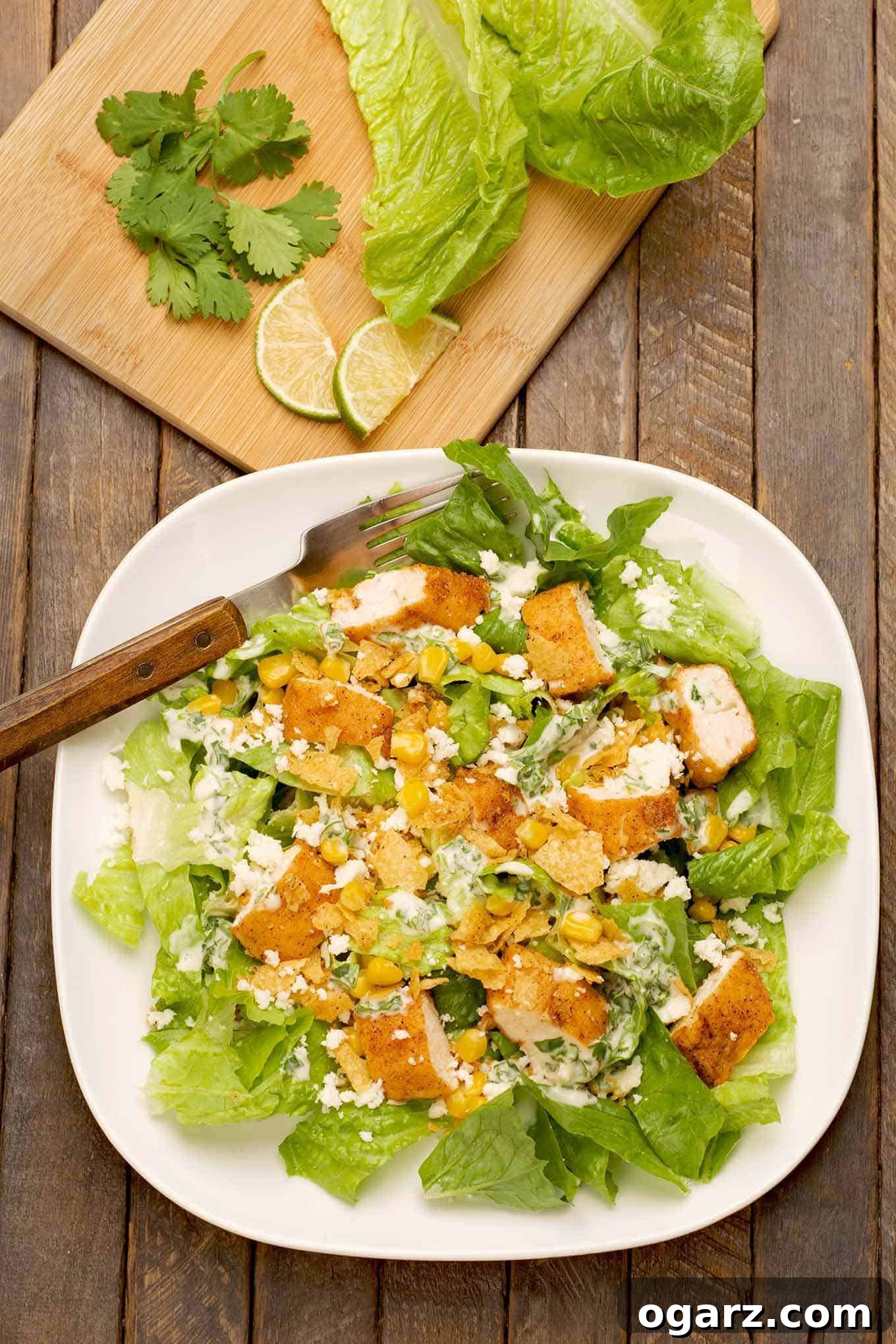 An overhead view of a vibrant Mexican-style Caesar salad; crisp romaine lettuce, perfectly seasoned chicken tenders, sweet corn kernels, crumbled queso fresco, and crunchy garlic-toasted tortilla chips, with fresh lime slices and cilantro in the background.