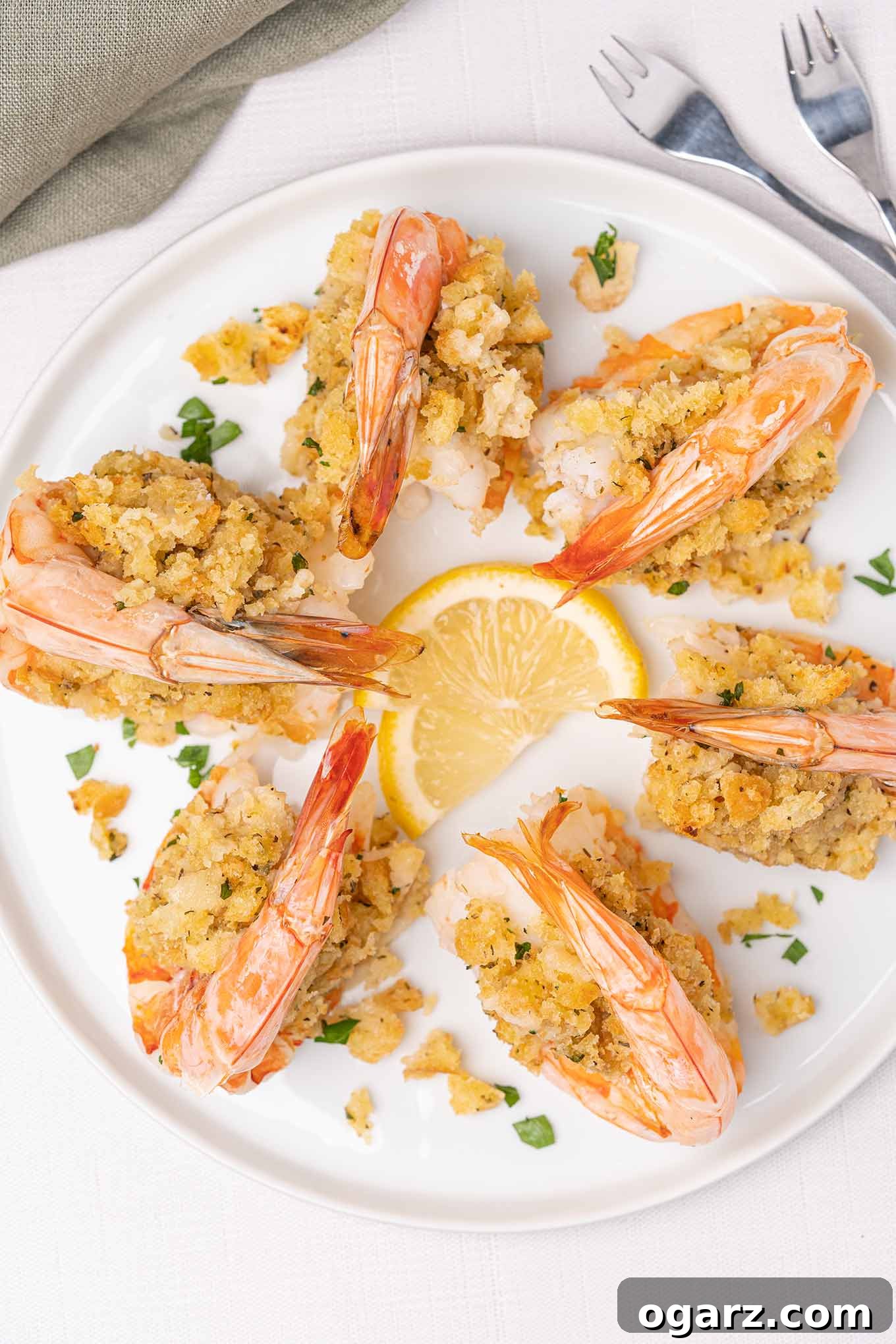 An overhead view of a round white plate filled with baked stuffed colossal shrimp with seafood forks and lemon slices for garnish, emphasizing its gourmet presentation.