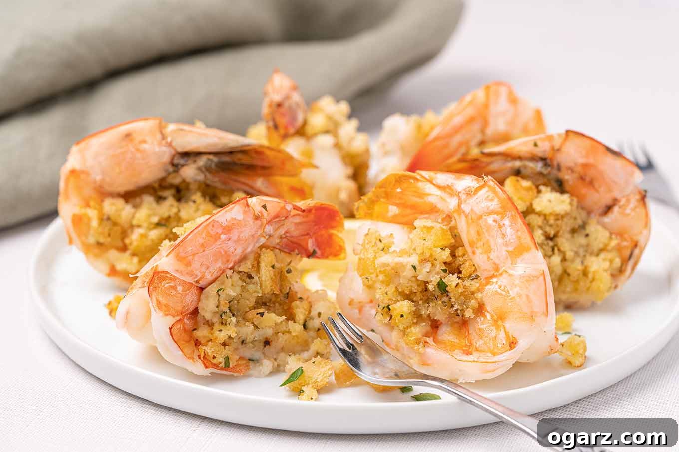 A side view of a serving of baked stuffed shrimp, showcasing the buttery, herb-infused fresh breadcrumb topping. A seafood fork rests elegantly on the white plate.