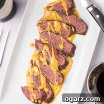 An overhead view of a white serving platter with slices of spice-crusted, roast venison tenderloin, drizzled with gjetost cheese sauce, a carving knife on the edge of the plate, and sprigs of fresh rosemary, peppercorns, and a glass of red wine off to the side.