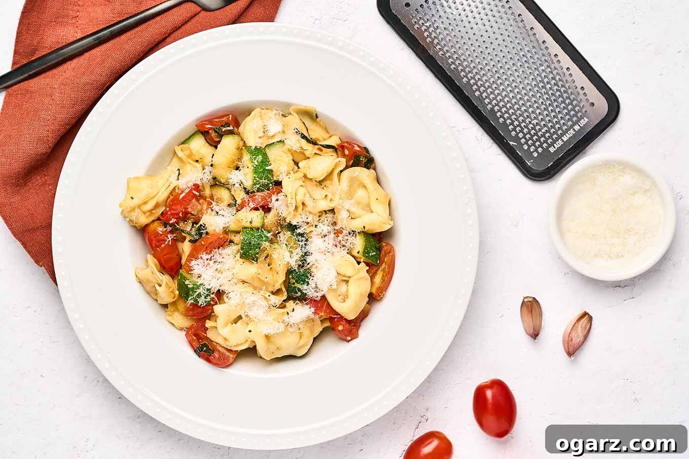Speedy Cheese Tortellini with Garden Vegetables 7 A perfectly plated serving of cheese tortellini with tender zucchini, sweet tomatoes, and fresh basil, artfully arranged in a bowl. The setting includes whole garlic cloves, ripe tomatoes, a small bowl of grated Parmesan, a crisp linen napkin, and a fork, highlighting the dish's fresh ingredients.