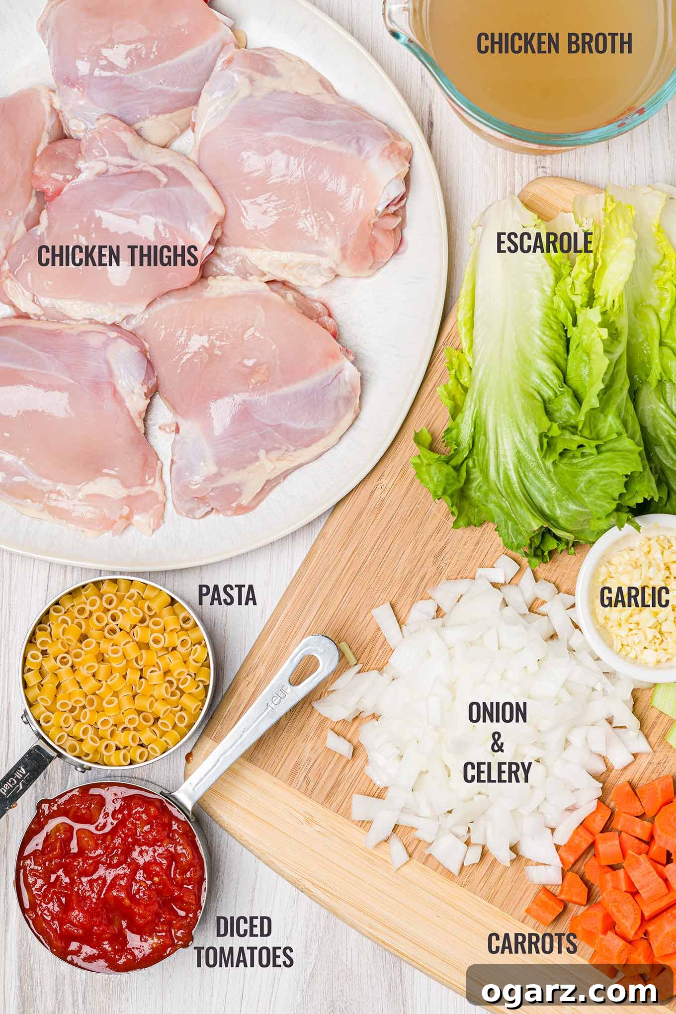 An overhead shot showcasing the fresh ingredients for Italian chicken soup, including boneless chicken thighs, chicken broth, vibrant escarole, chopped onion, sliced celery, minced garlic, crisp carrots, diced tomatoes, and ditalini pasta.