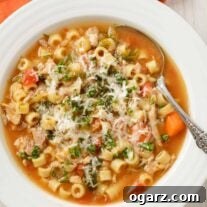 A bowl of Italian chicken soup with ditalini, carrots, escarole, and chicken, sprinkled with grated parmesan cheese and chopped fresh parsley.