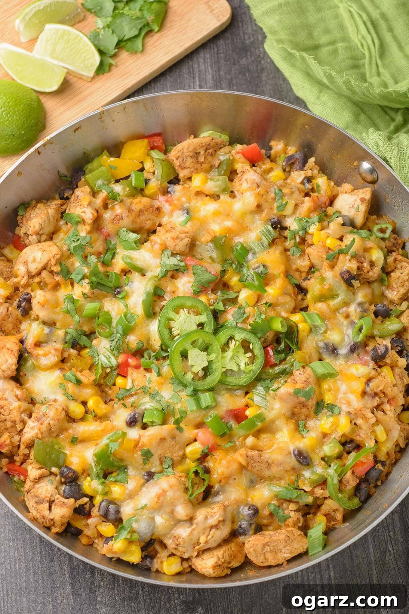 Fiesta Chicken Cauliflower Bowl 2 A vibrant frying pan filled with Southwest-style chicken and cauliflower rice, adorned with colorful bell peppers, sweet corn, fresh scallions, hearty black beans, and garnished with spicy sliced jalapenos and fragrant cilantro.