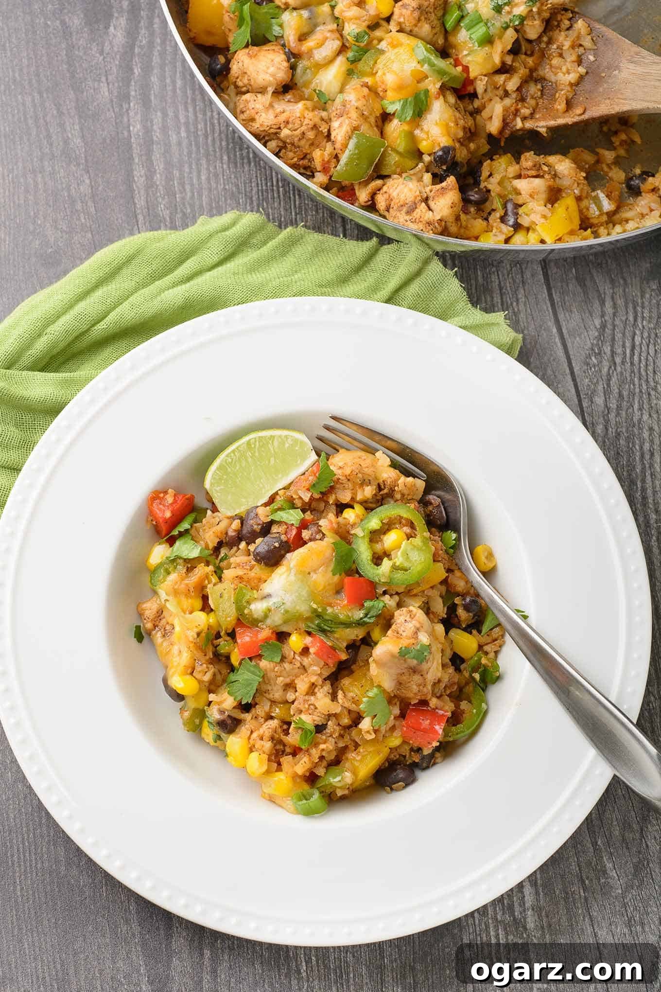 Fiesta Chicken Cauliflower Bowl 3 A perfectly plated serving of Southwest chicken and cauliflower rice in a shallow bowl, garnished with a vibrant lime wedge and a spicy jalapeño slice. The cooking skillet, still warm and inviting, sits in the background.
