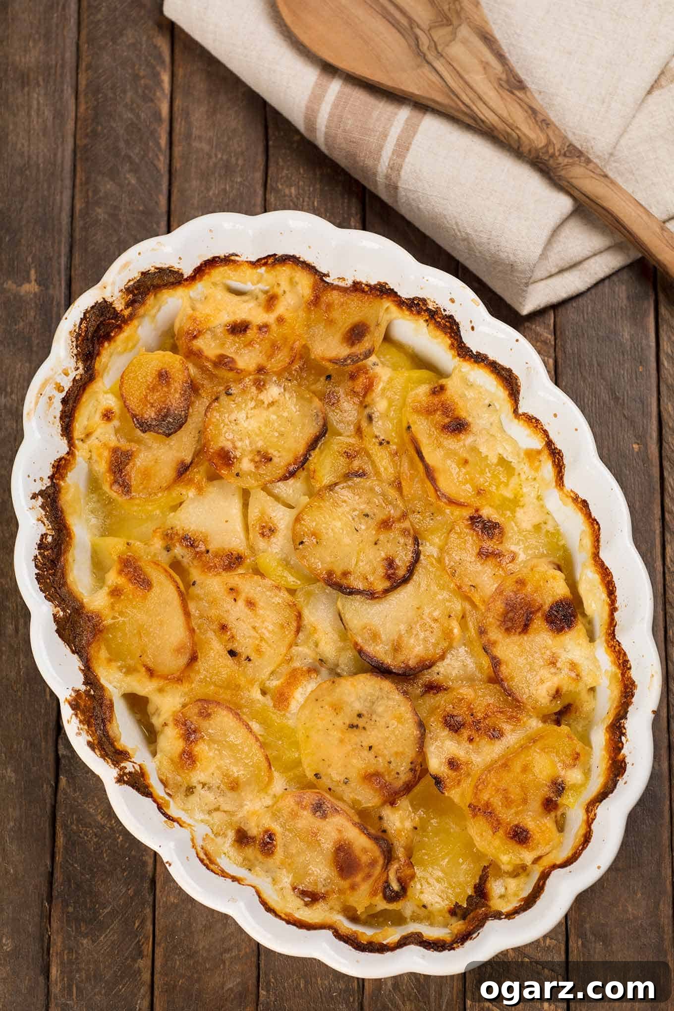 Creamy Potato Gratin 2 An oval casserole of golden-brown scalloped potatoes, with a wooden spoon and tea towel in the background, signaling warmth and comfort.