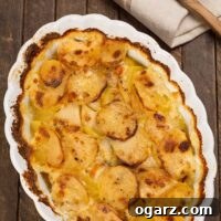 An oval casserole of scalloped potatoes, browned on the top, with a wooden spoon and tea towel in the background.