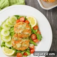 A composed green salad with lettuce, cherry tomatoes, and cucumber slices topped with lemon slices, fresh dill, two salmon cakes, and dill dressing.