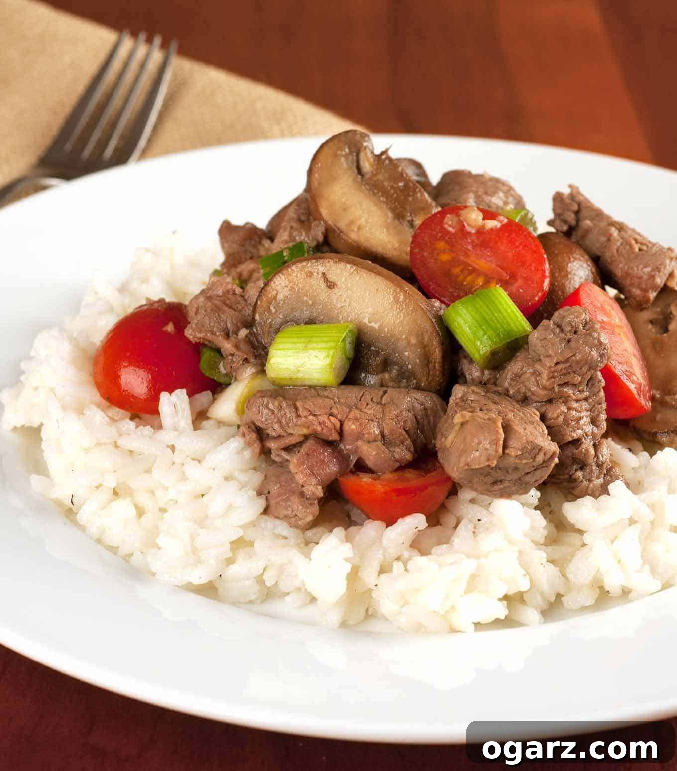 Succulent Garlic Mushroom Tenderloin Morsels 2 Tenderloin beef tips sautéed with mushrooms, cherry tomatoes, and scallions served over a bed of white rice on a white plate.