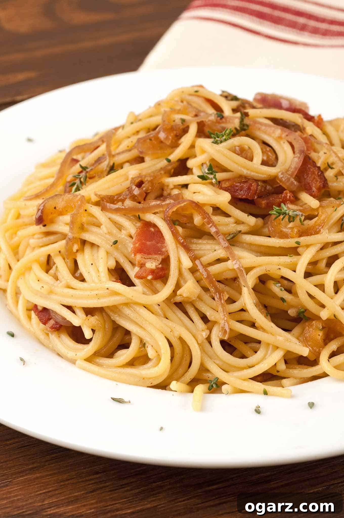 Spaghetti tossed with crispy bacon, caramelized onions, and fresh thyme on a white dish, a striped towel in the background.