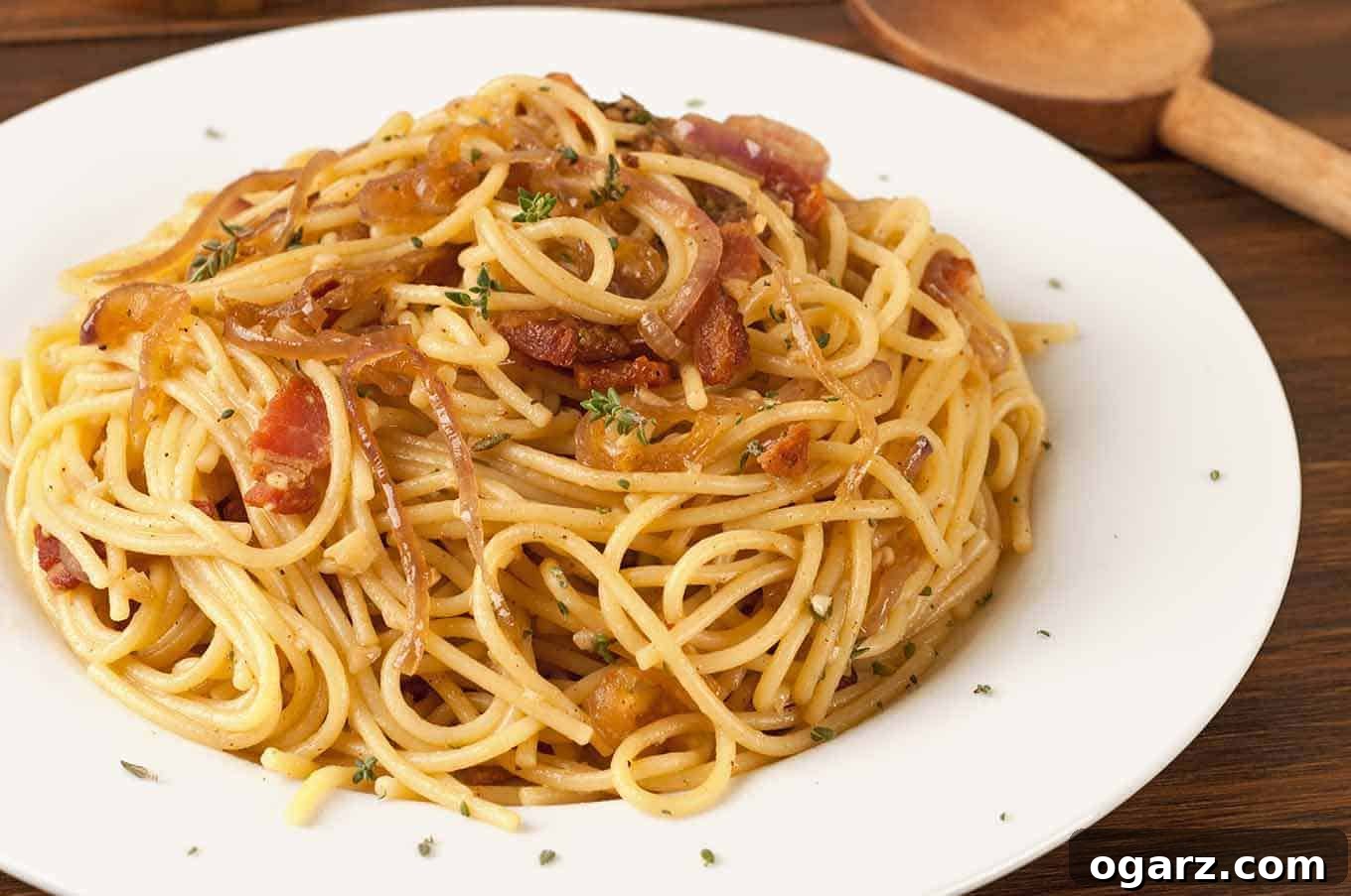 A plate of spaghetti tossed with crispy bacon, caramelized onions, and fresh thyme on a white dish.