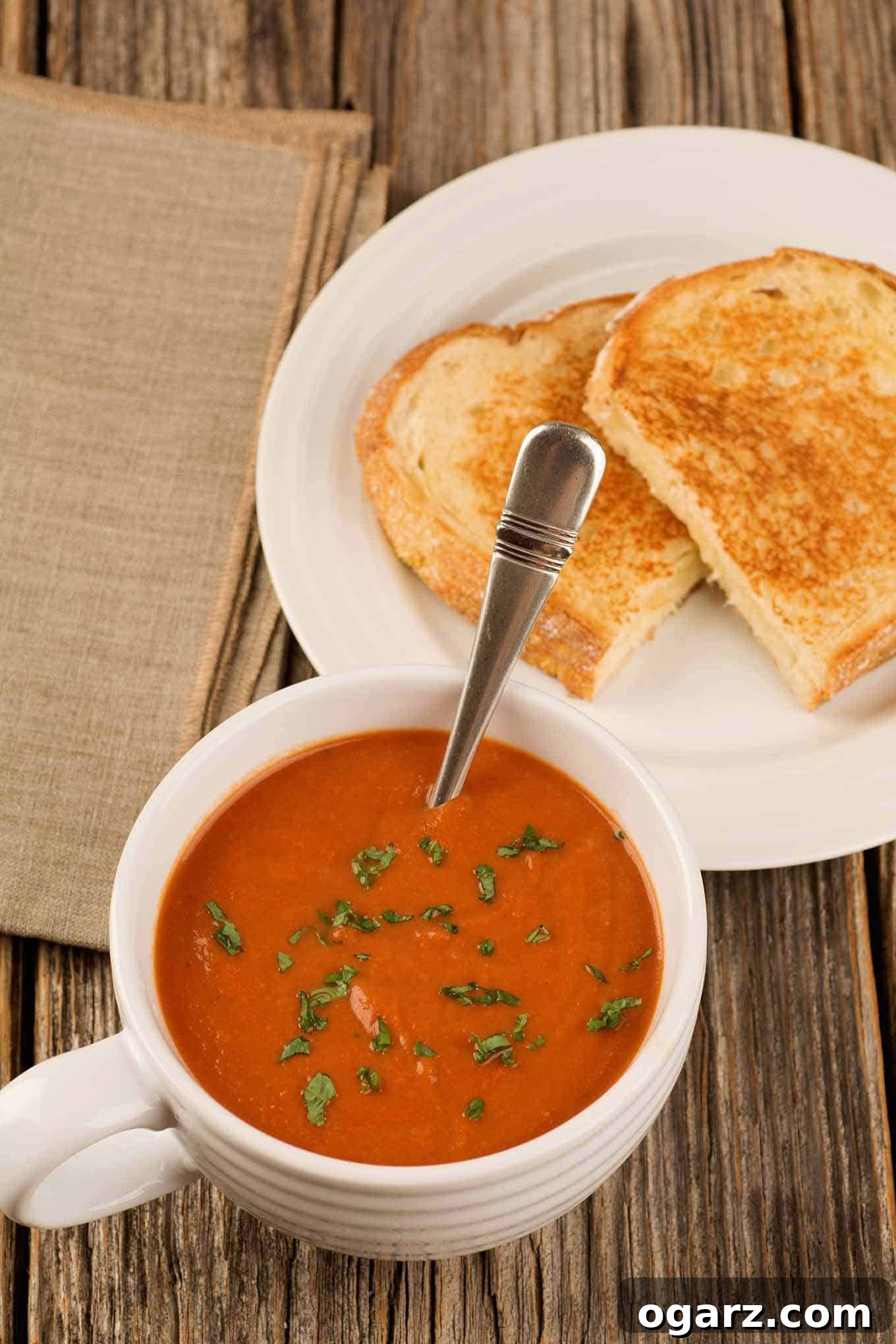 A large cup of tomato basil soup garnished with fresh basil next to a plate with a grilled cheese sandwich and a napkin.