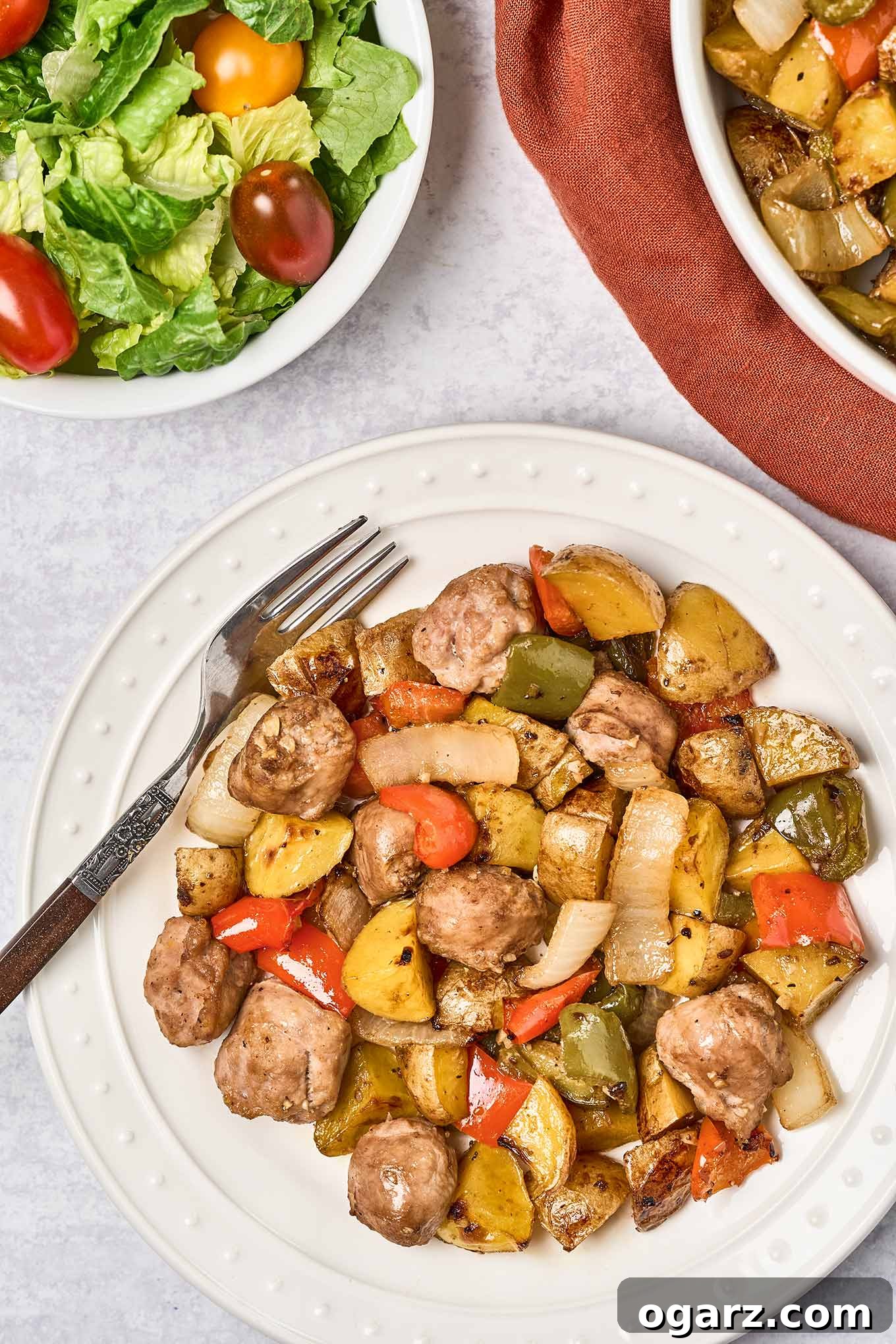 Hearty Sausage, Pepper, and Potato Roast 2 A round white dinner plate with balsamic glazed baked Italian sausage, bell peppers, and onions, a dinner fork to the side and a serving bowl with more of the main dish and a bowl of salad with grape tomatoes in the background.