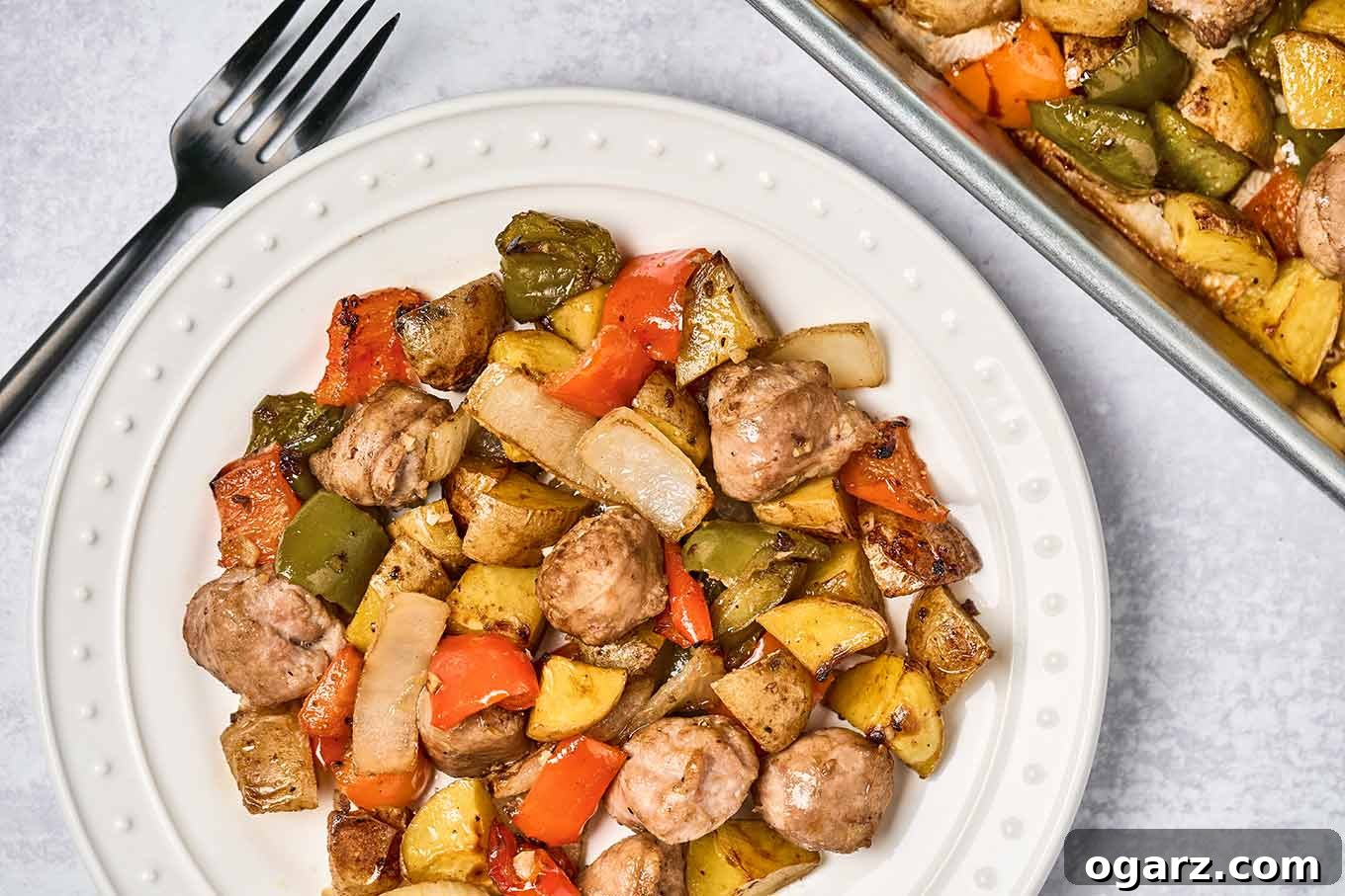 Hearty Sausage, Pepper, and Potato Roast 4 A single serving of baked Italian sausage with bell peppers, potatoes, and onions on a round white plate with the sheet pan it was served from in the upper right, dinner fork on the left.