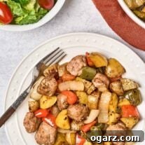A round white dinner plate with balsamic glazed baked Italian sausage, bell peppers, and onions, a dinner fork to the side and a serving bowl with more of the main dish and a bowl of salad with grape tomatoes in the background.