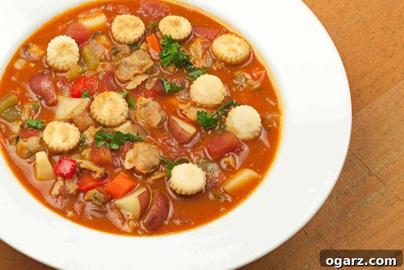 A vibrant bowl of Manhattan Clam Chowder, rich with tomatoes, fresh clams, bacon, and colorful vegetables, garnished with parsley and oyster crackers.