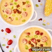 An overhead view of two white soup crocks of corn chowder garnished with bacon, tomatoes, corn, and scallions, a whole ear of corn, a spoon, and a scattering ingredients in the background.