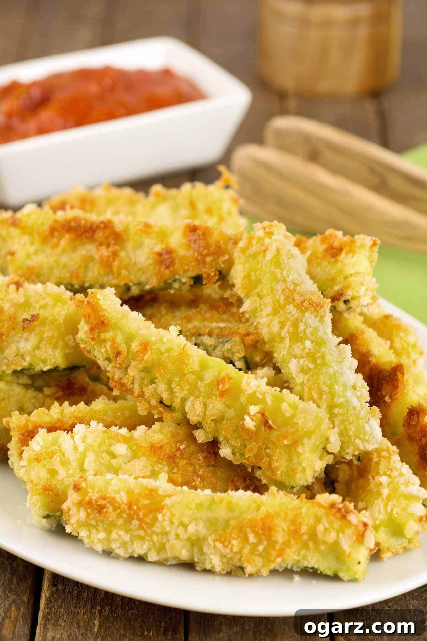 Crispy oven-fried zucchini sticks coated in panko crumbs and grated Parmesan stacked on a serving plate with marinara dipping sauce in the background.