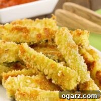 Crispy oven-fried zucchini sticks coated in panko crumbs and grated Parmesan stacked on a serving plate with marinara dipping sauce in the background.