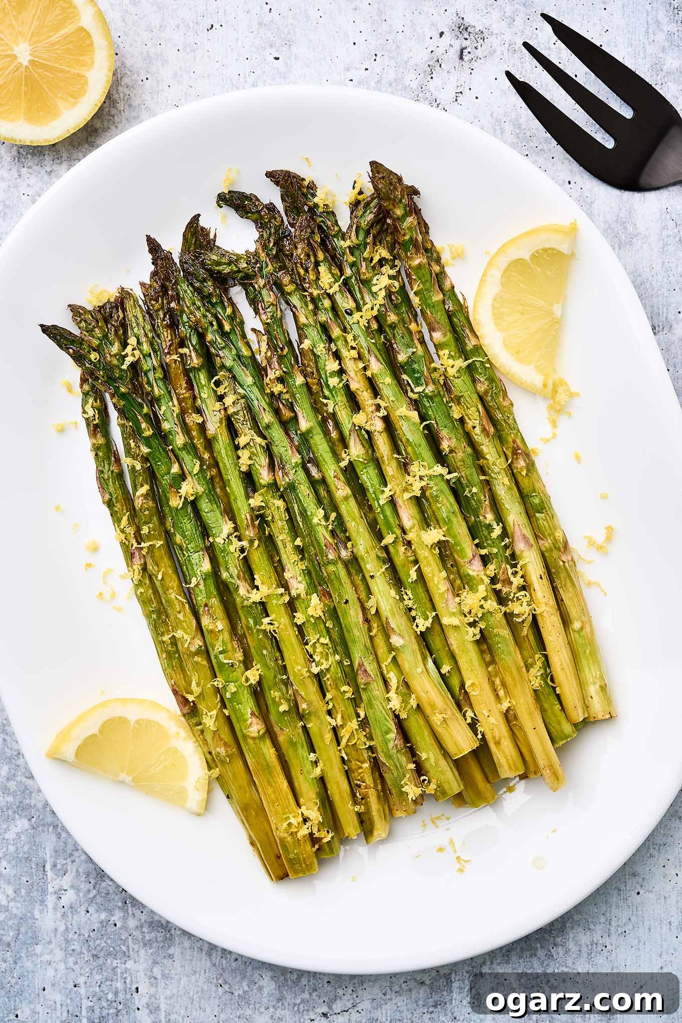Zesty Roasted Asparagus 2 A serving plate of lemon roasted asparagus topped with lemon zest and garnished with lemon slices, a black serving fork an half a lemon in the background.