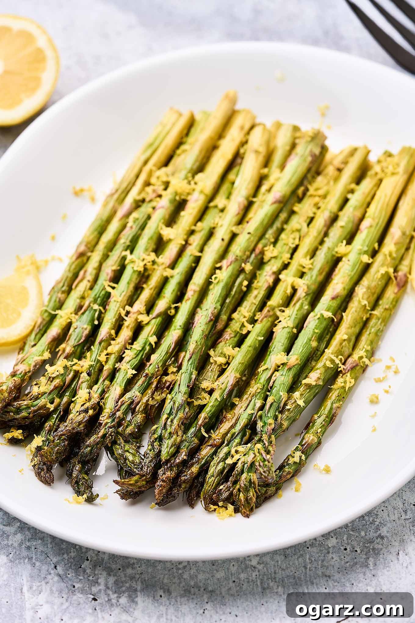 Zesty Roasted Asparagus 6 Spears of lemon roasted asparagus topped with lemon zest on a white oval serving plate garnished with lemon slices.