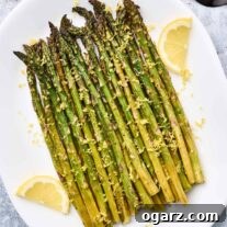 A serving plate of lemon roasted asparagus topped with lemon zest and garnished with lemon slices, a black serving fork an half a lemon in the background.