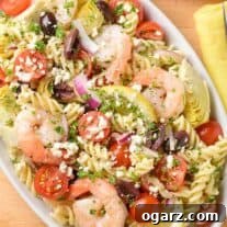 An overhead view of Mediterranean pasta salad with shrimp, artichoke hearts, Kalamata olives, cherry tomatoes, and feta cheese, extra lemon in the background.