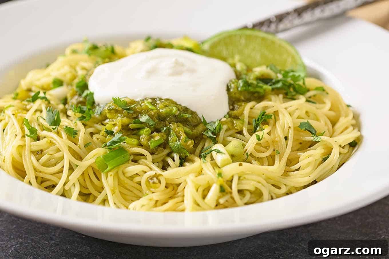 A close-up view of thin spaghetti luxuriously tossed with a bright green salsa verde, featuring roasted tomatillos, poblano peppers, lime, scallions, and cilantro, elegantly crowned with a dollop of sour cream.