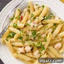 A plate of penne pasta tossed with tender lobster, shallots, white wine, cream, butter, and a drizzle of truffle oil, garnished with chopped parsley.
