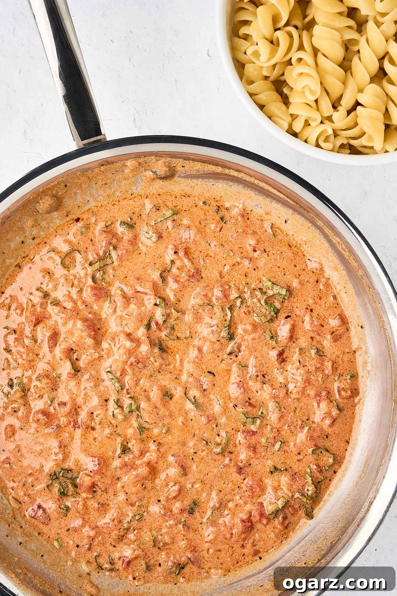 A pan filled with creamy mascarpone tomato sauce with fresh basil next to a bowl of cooked fusilli pasta, ready to be combined.
