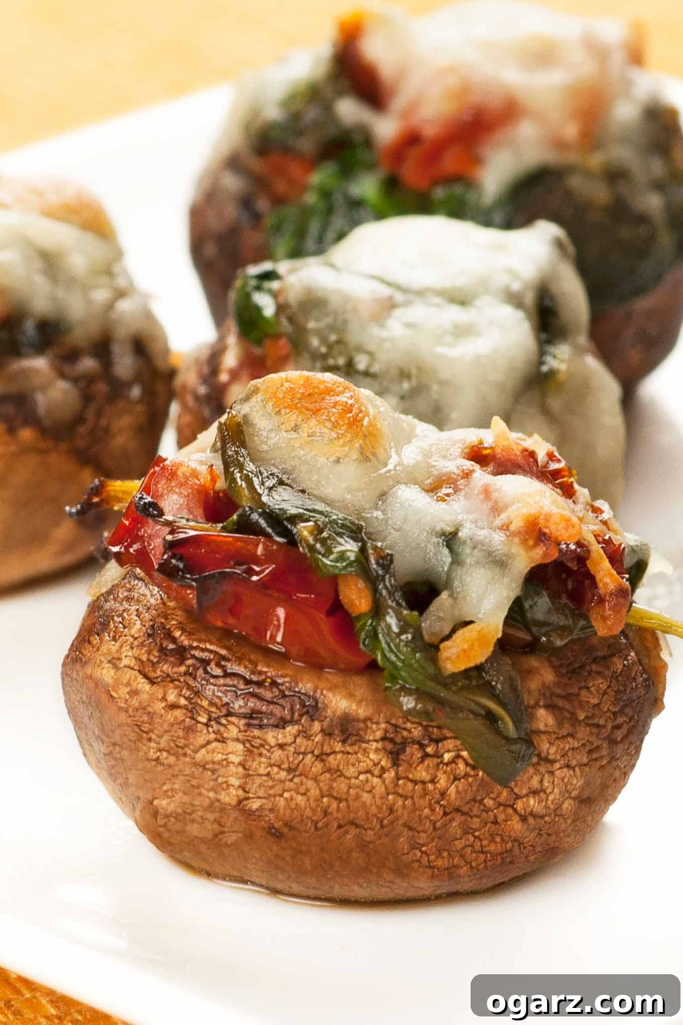Close-up photo of spinach stuffed mushrooms with melted Asiago cheese and sun-dried tomatoes on top, served on a white plate. A perfect bite-sized appetizer.