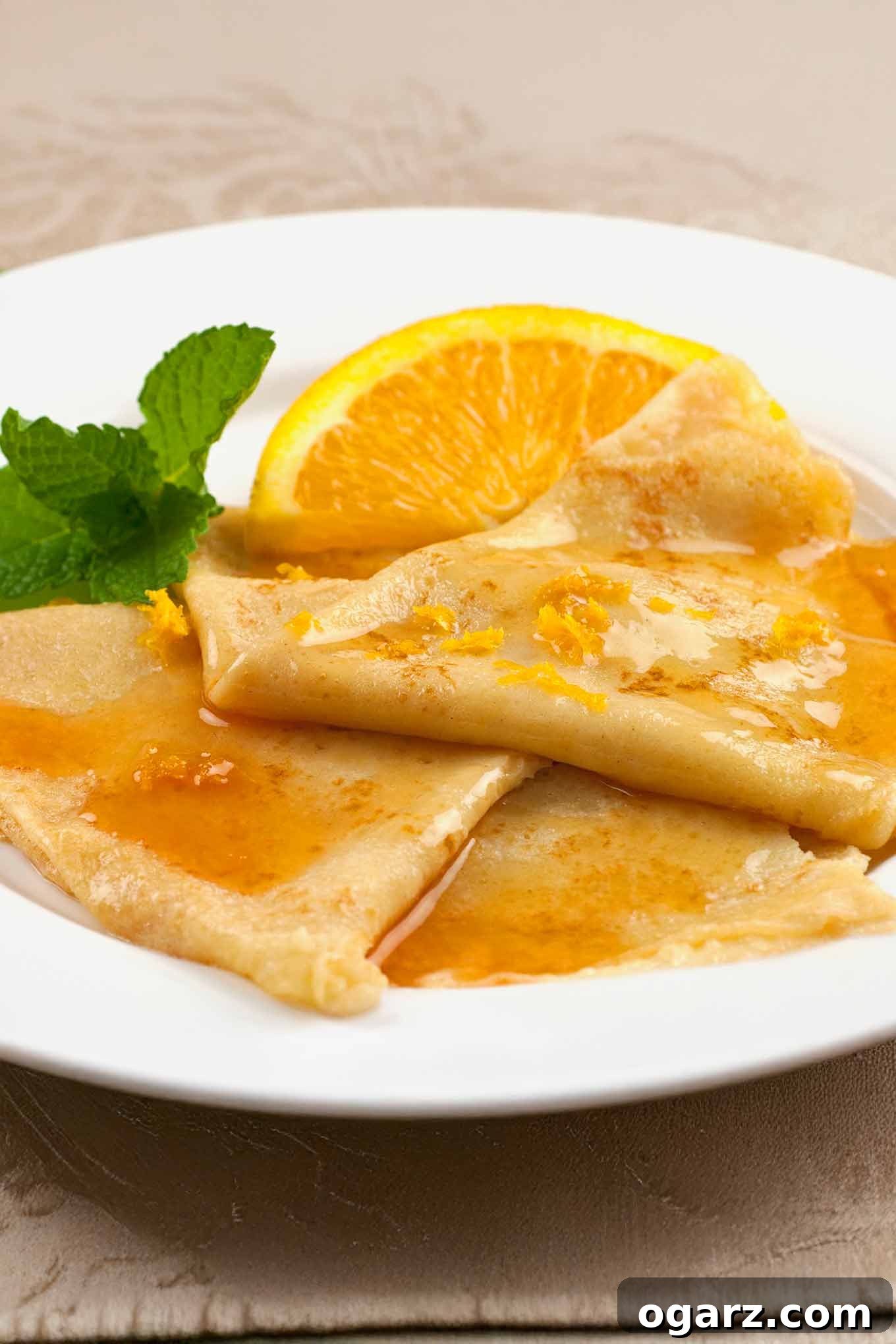 Three Crêpes Suzette on a round white plate, topped with Grand Marnier sauce and orange zest, and garnished with a sprig of mint and a fresh orange wedge.