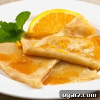 Three Crêpes Suzette on a round white plate, topped with Grand Marnier sauce and orange zest, and garnished with a sprig of mint and a fresh orange wedge.