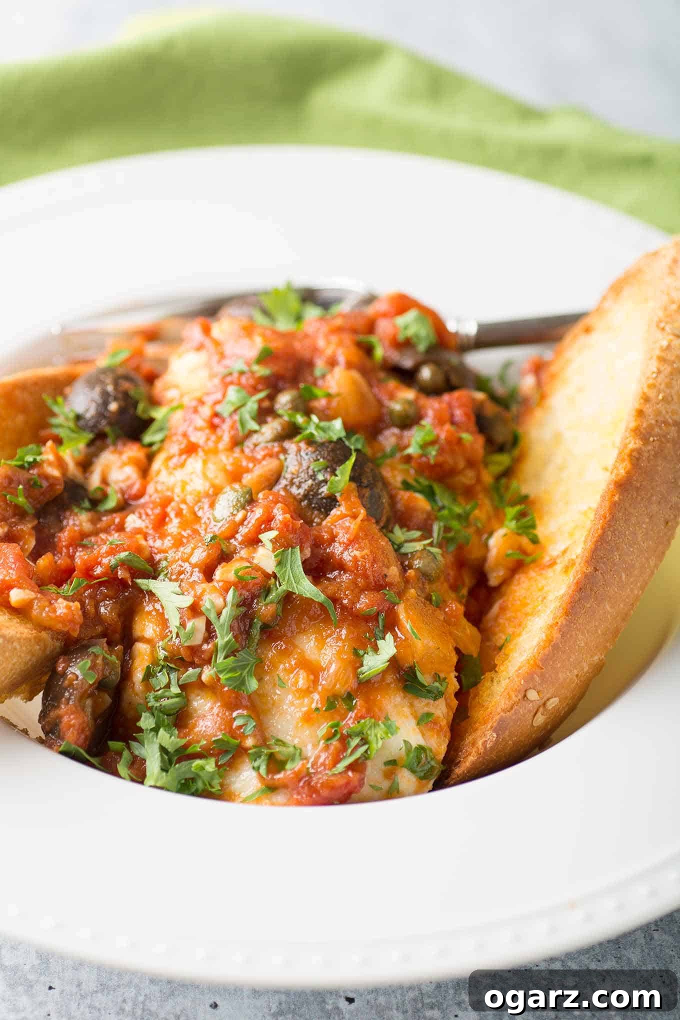 A shallow bowl with a fish fillet topped with puttanesca tomato sauce made with garlic, onion, and kalamata olives, and two slices of garlic toast. 
