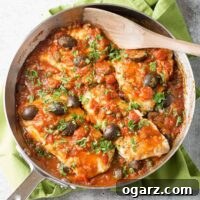 An overhead view of a frying pan filled with white fish fillets in puttanesca-style tomato sauce with kalamata olives, garnished with parsley.